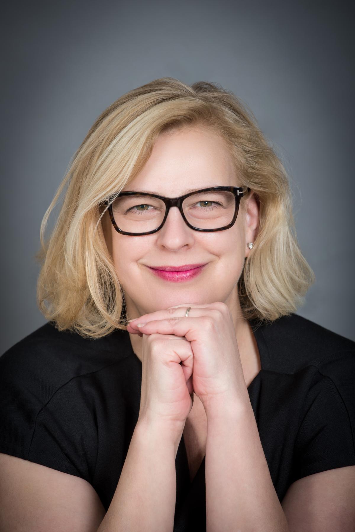Julie Garcia, a blonde woman with glasses, smiles at the camera with her hands folded under her chin.